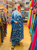 A woman takes a mirror selfie in a clothing store wearing a blue floral print salwar kameez set with a matching dupatta. The background is filled with racks of colorful garments and shelves of folded fabrics.