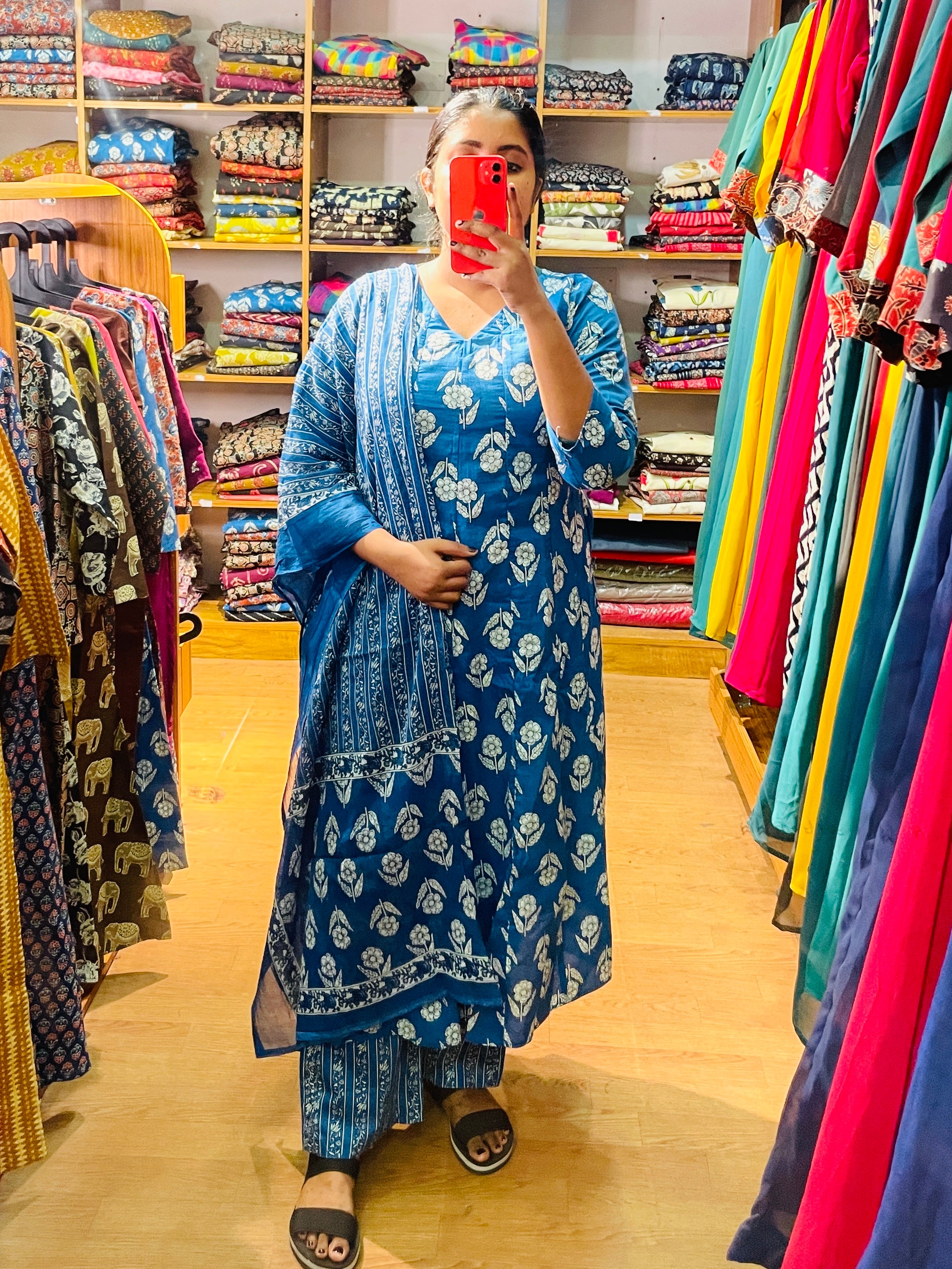 A woman takes a mirror selfie in a clothing store wearing a blue floral print salwar kameez set with a matching dupatta. The background is filled with racks of colorful garments and shelves of folded fabrics.