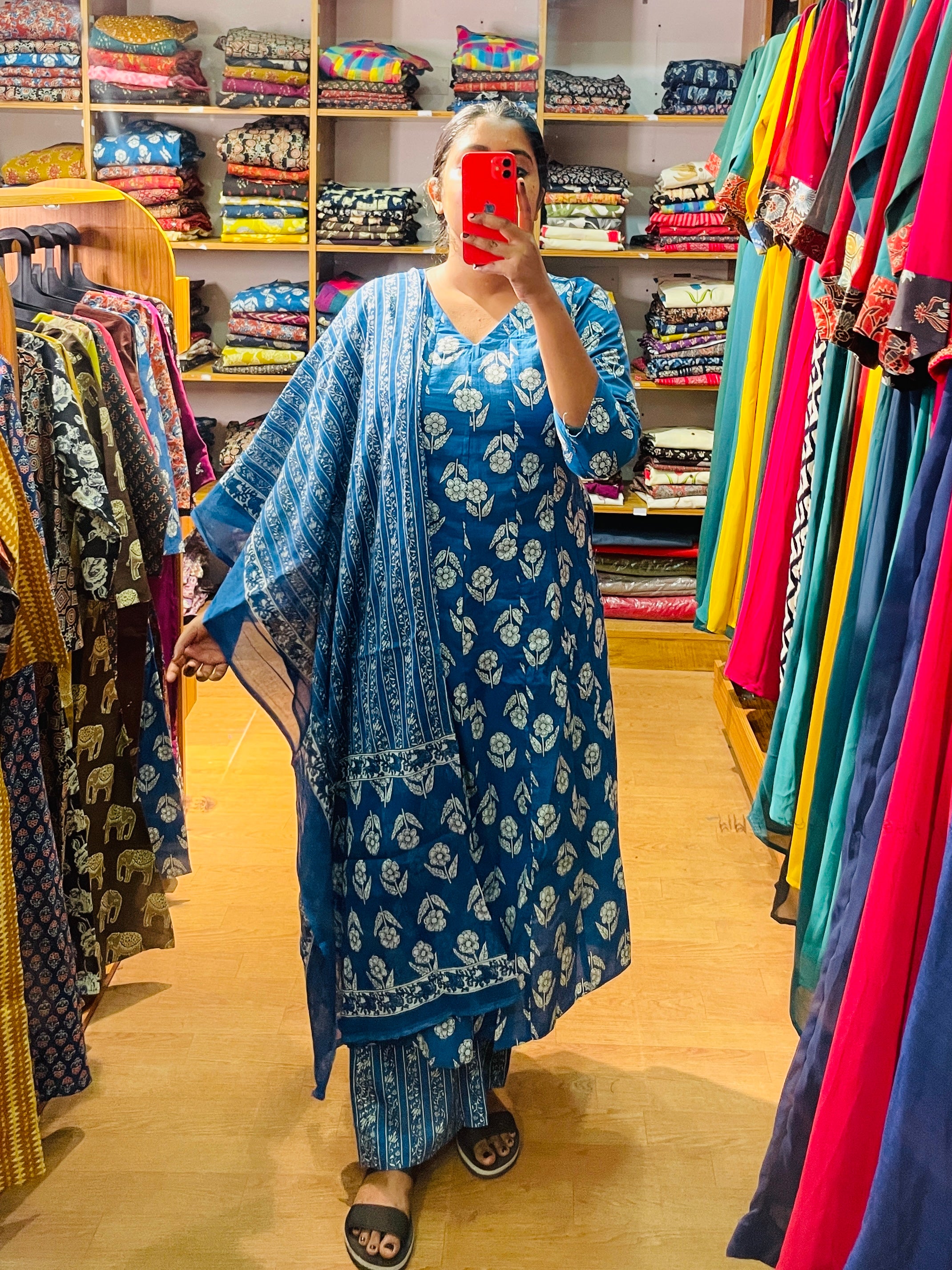 A woman taking a mirror selfie in a clothing store wearing a blue block-printed kurta set with a matching dupatta. The set features white floral block prints and is displayed against a background of colorful clothing racks and shelves.